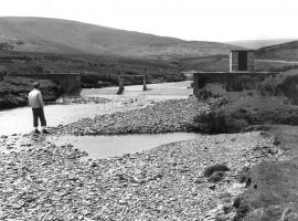 A historical black and white photo of a gauging station. A man is standing downstream of the weir. The river is in a state of low flows.Gauging Station